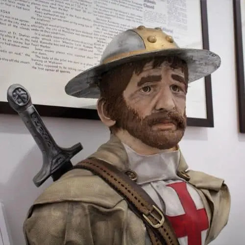 Busto de um cavaleiro medieval com túnica de cruz vermelha, chapéu de metal e espada nas costas